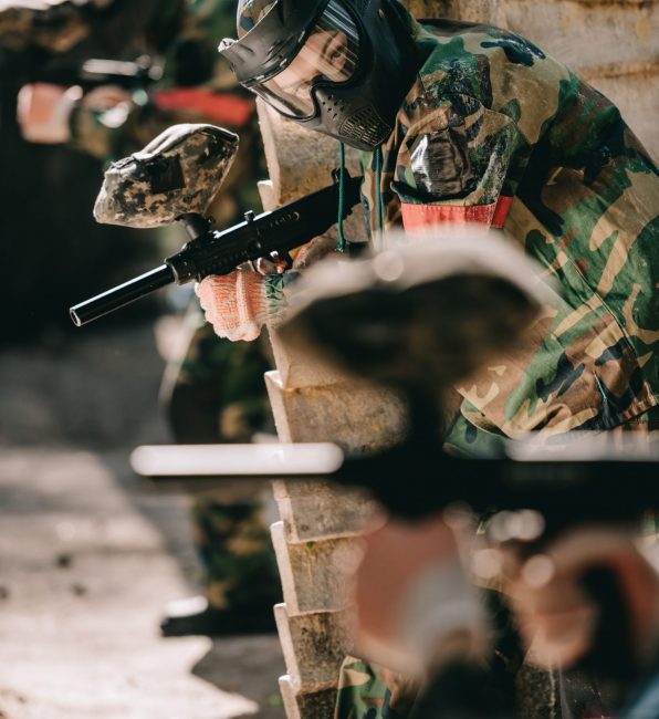 selective focus of female paintball player in camouflage and protective mask holding marker gun outdoors